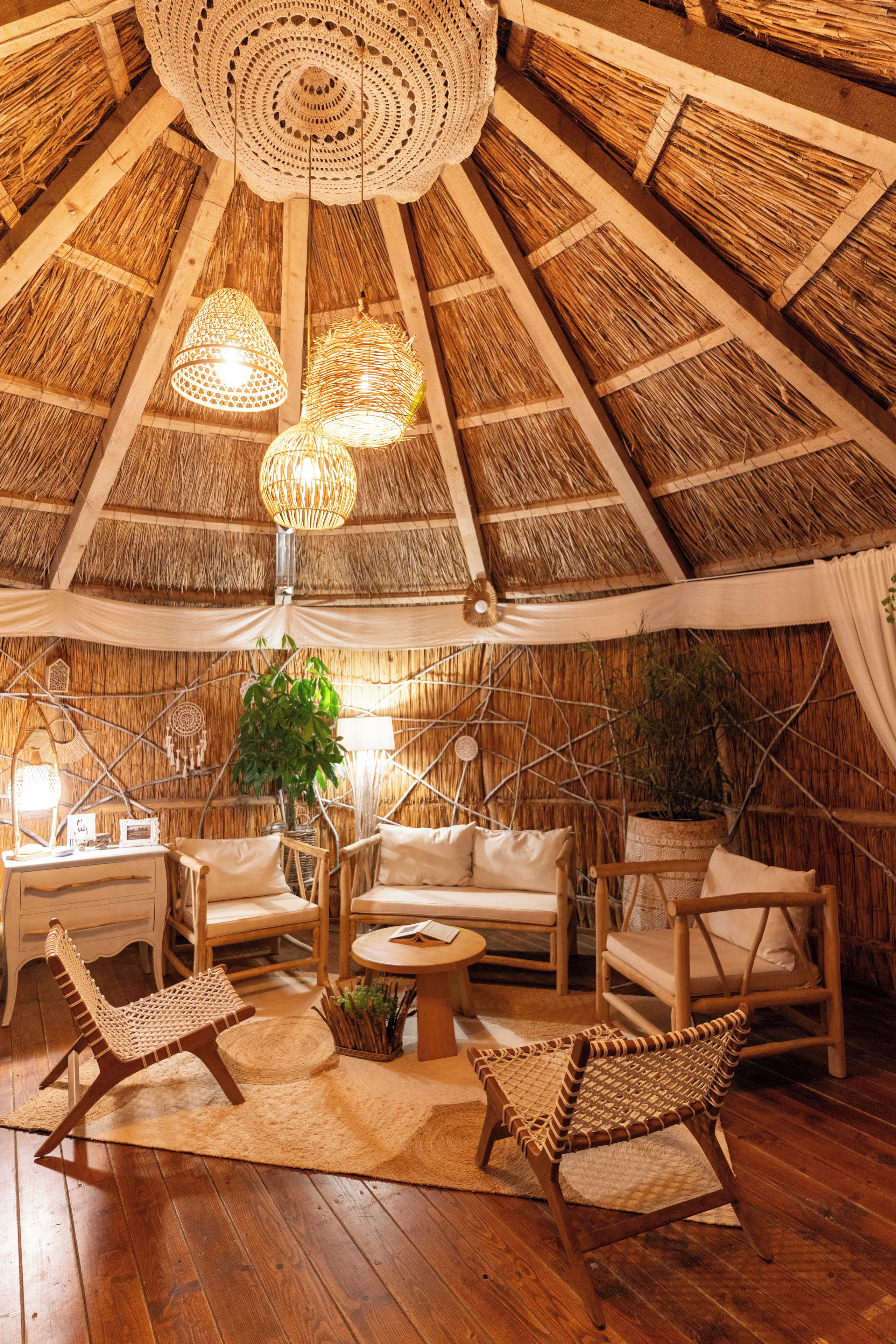 Una habitación con techo de paja, muebles de mimbre, una lámpara colgante tejida y una planta en el centro.