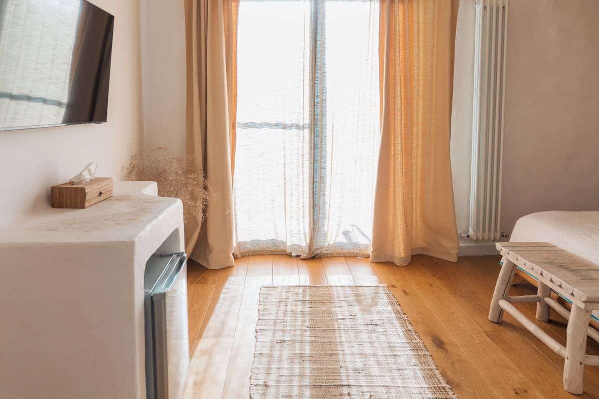 A modern bedroom with a large window, a wooden floor, a white bed, a small bedside table, and a hanging light fixture.