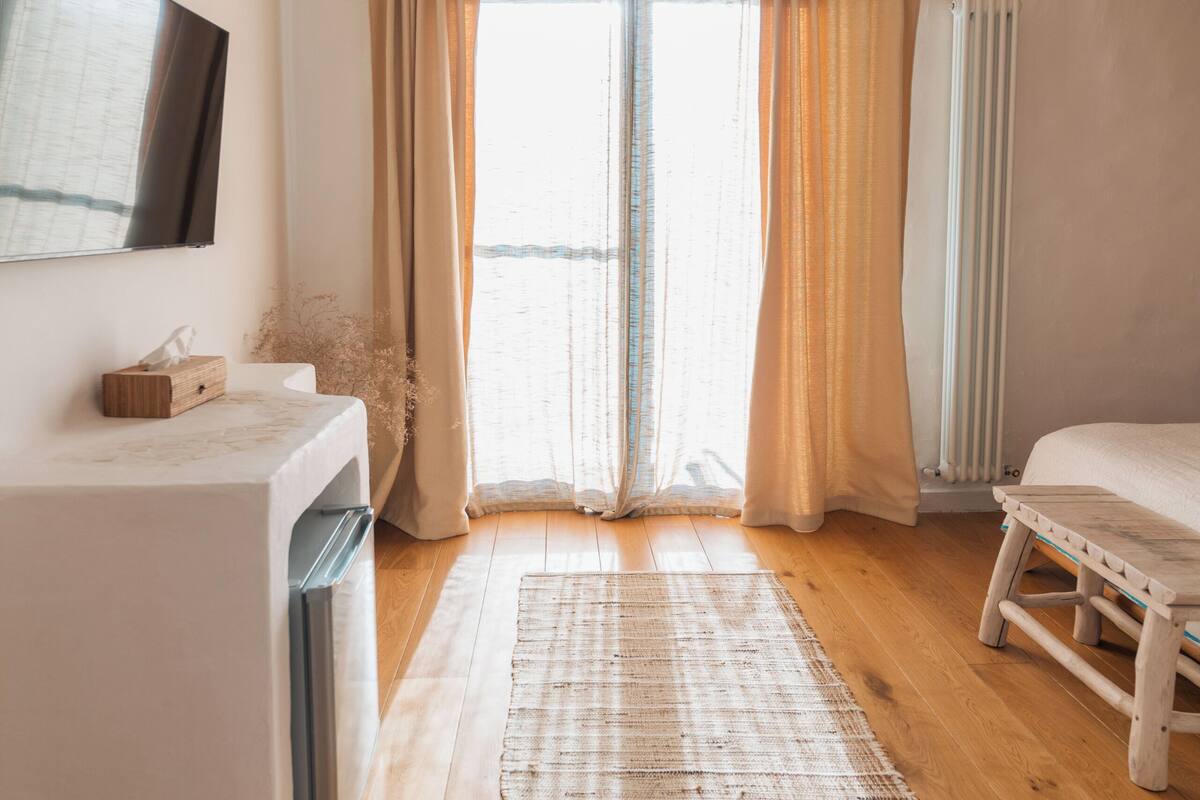 Ein modernes Schlafzimmer mit großem Fenster, Holzboden, weißem Bett, einem kleinen Nachttisch und einer Pendelleuchte.