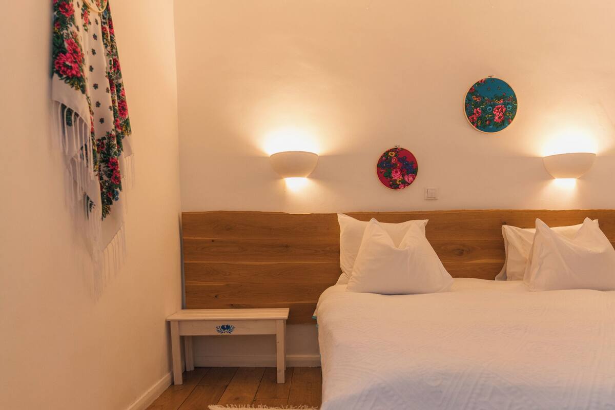 A bedroom with a wooden headboard, a white bedspread, two wall-mounted lights, a small bedside table, and a decorative wall hanging.