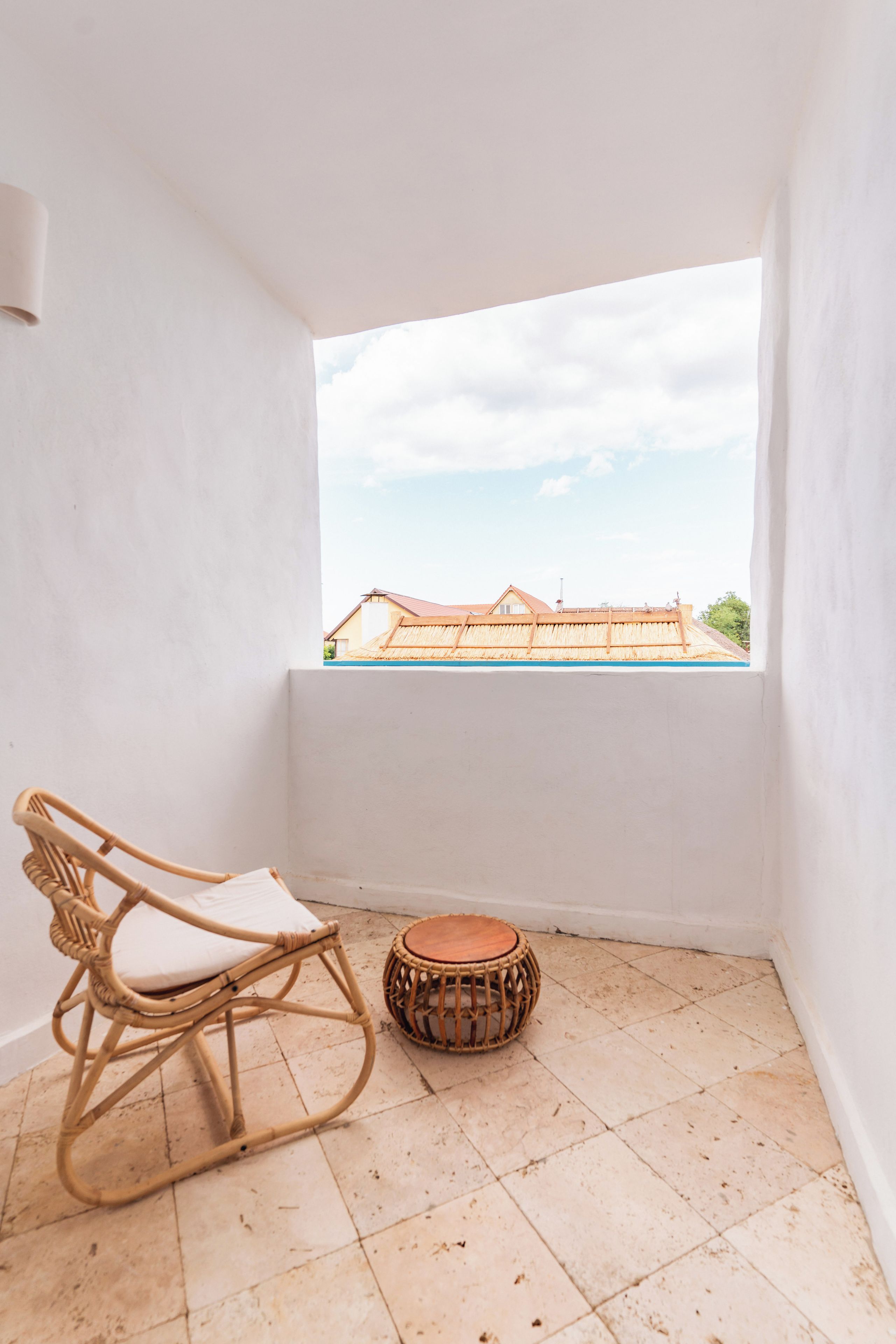 A balcony with a wicker rocking chair and a drum, a view of a building with a red roof, and a cloudy sky.