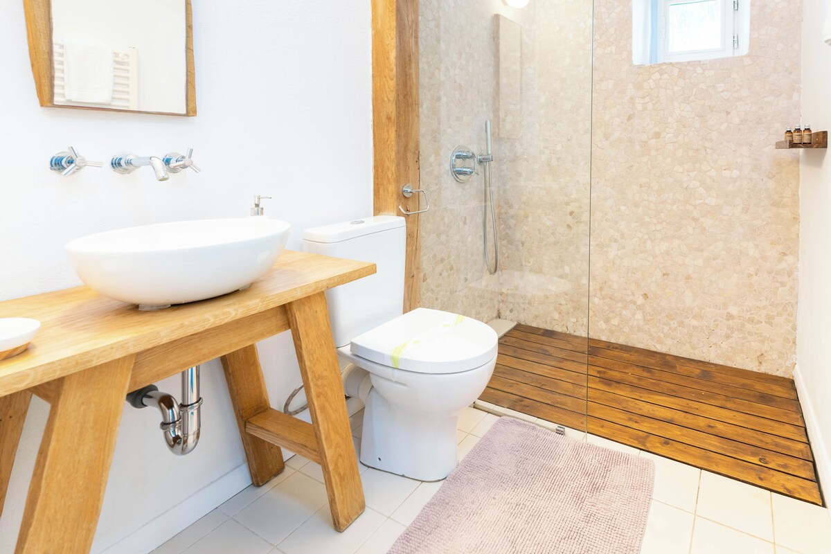A modern bathroom with a wooden vanity, a large mirror, a white sink, a toilet, and a glass-enclosed shower.