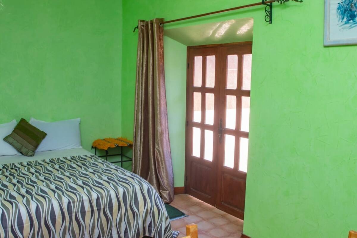 A room with a bed, a table, a chair, a door, and an air conditioning unit.