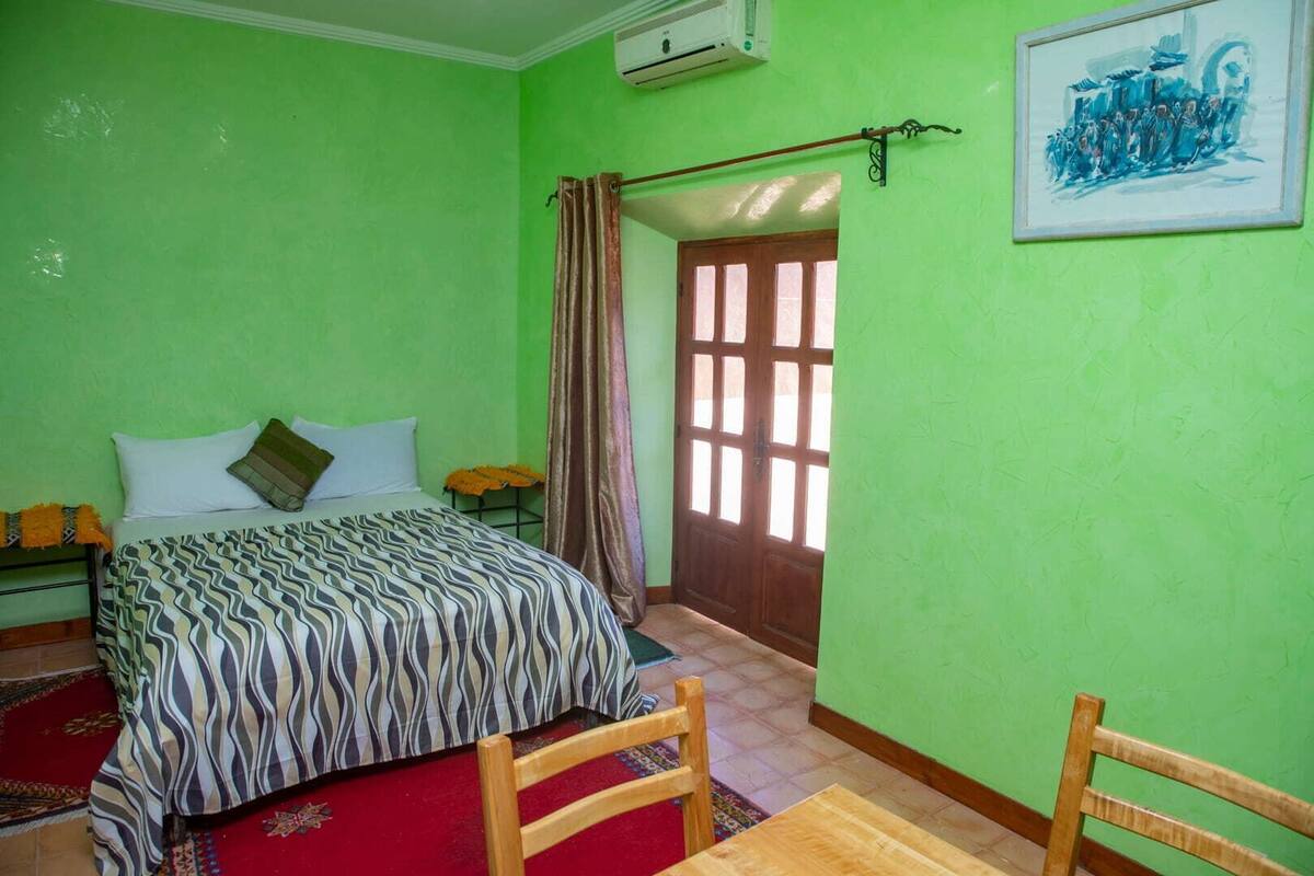 A room with a bed, a wooden chair, a table, a door, and a painting on the wall.