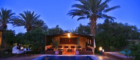 A poolside area with a covered seating area, palm trees, and a clear sky.