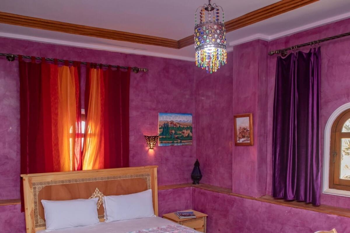 A bedroom with a wooden bed, a nightstand, a chair, a window with curtains, and a chandelier.