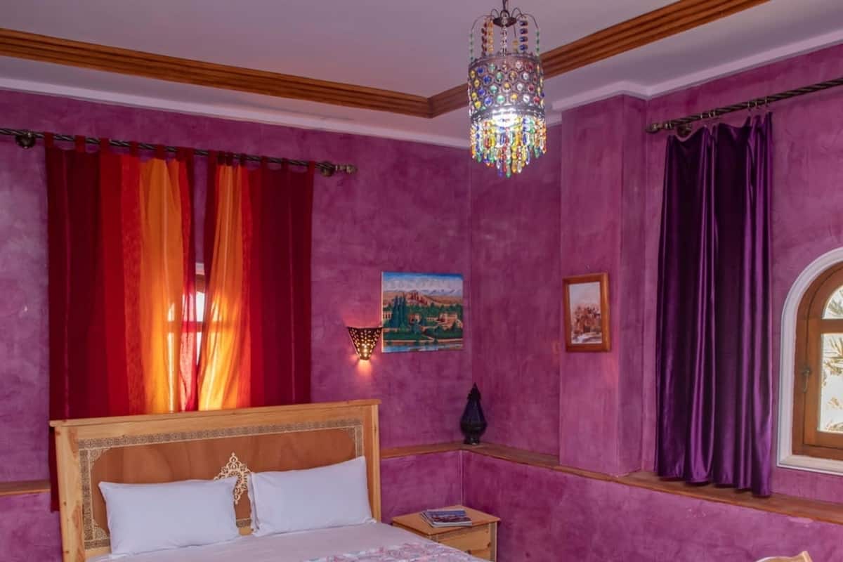 A bedroom with a wooden bed, a nightstand, a chair, a window with curtains, and a chandelier.