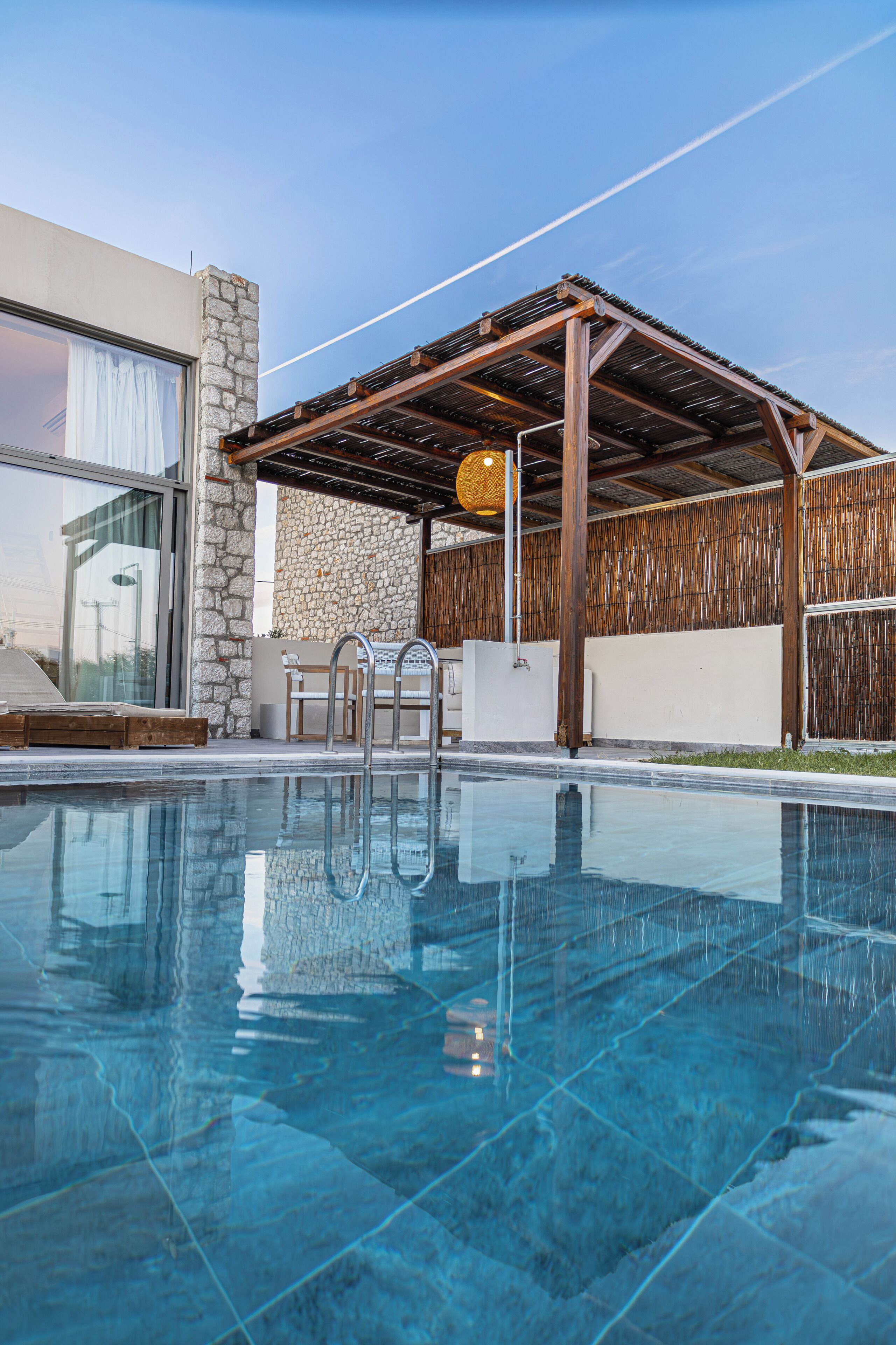 A swimming pool with a stone wall, wooden pergola, and a glass door.