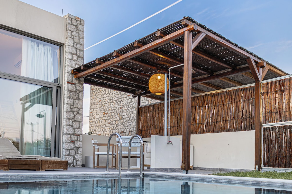 Une piscine bordée d’un mur en pierre, agrémentée d’une pergola en bois et surplombée d’un bâtiment moderne doté de grandes fenêtres.