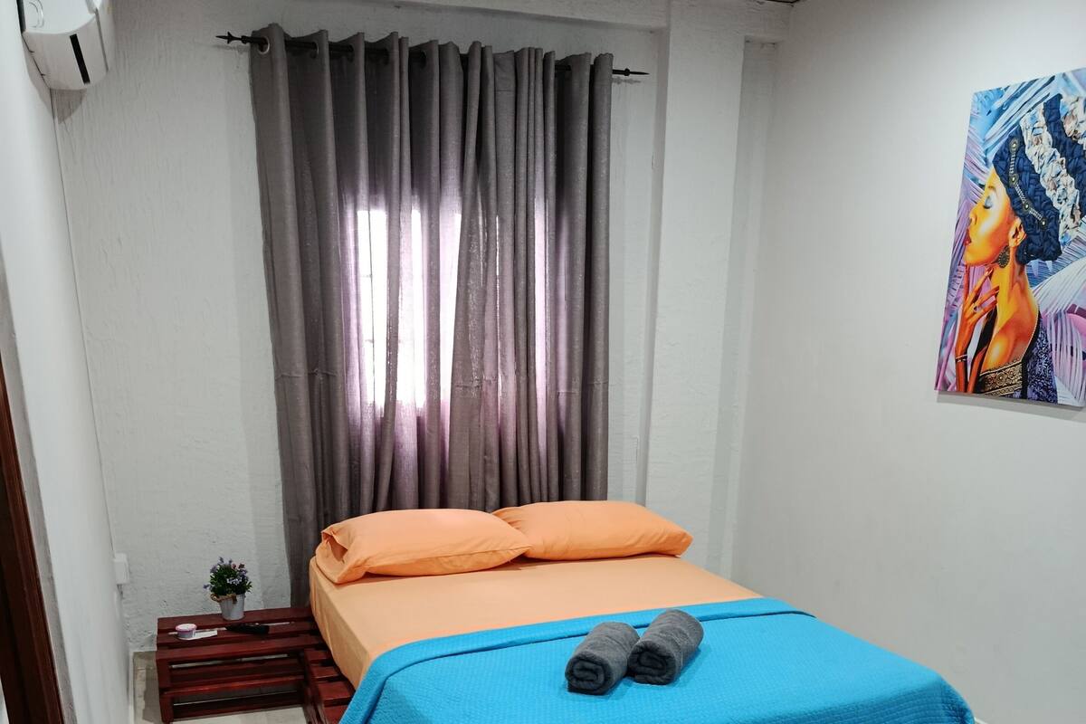 A small, neatly arranged bedroom with a bed, a chair, a small table, and a potted plant.