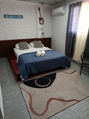 A neatly made bed with a blue bedspread and two pillows, a wall clock, and a guitar hanging on the wall. - Hostal Cigarra (Panama City)