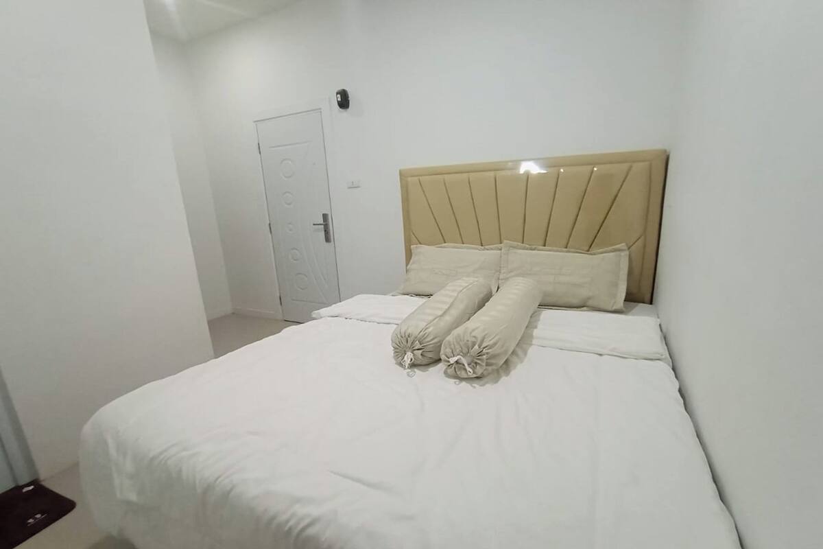 A neatly made bed with a beige headboard and white bedding in a simple, clean room with white walls and a door in the background.