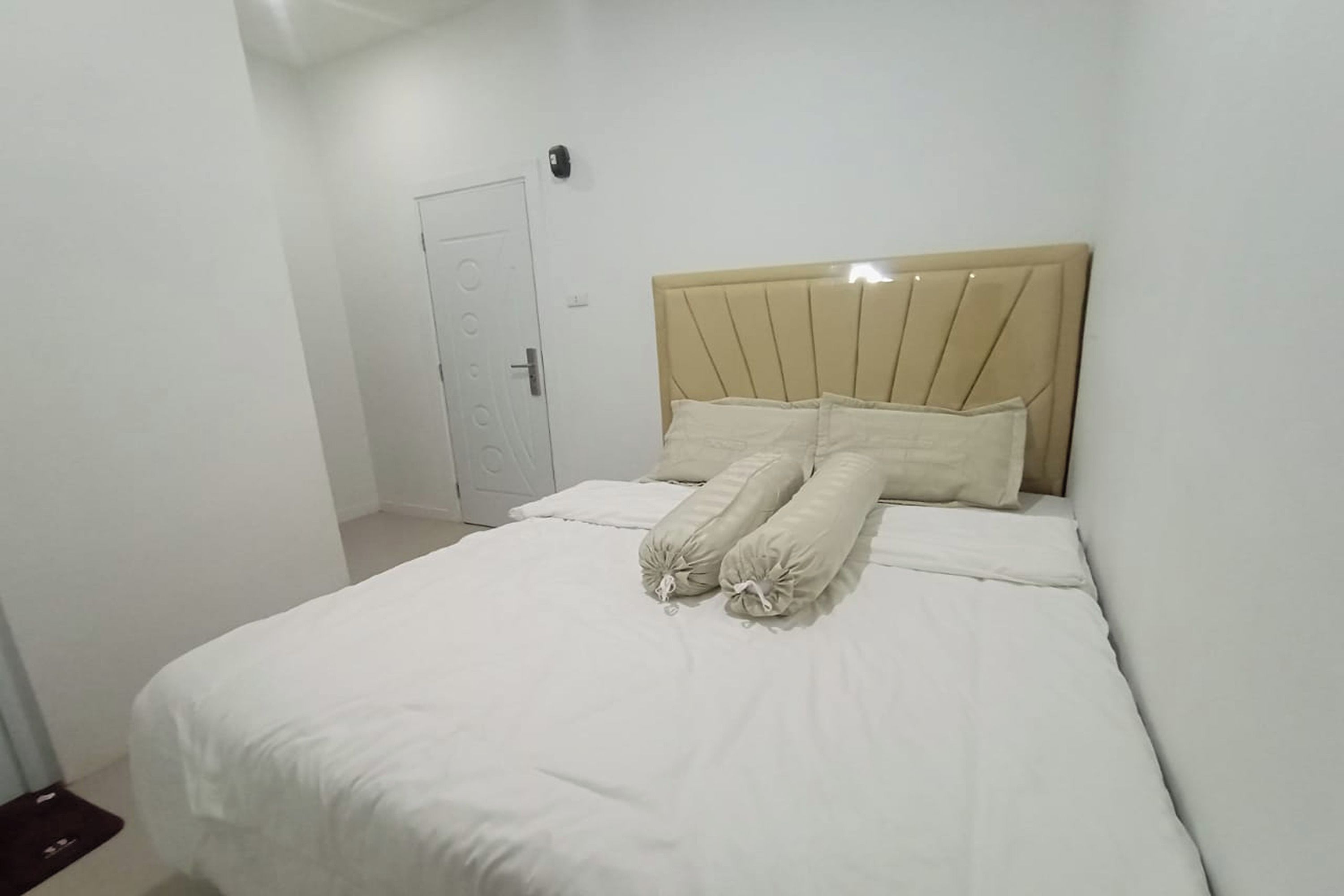A neatly made bed with a beige headboard and white bedding in a simple, clean room with white walls and a door in the background.