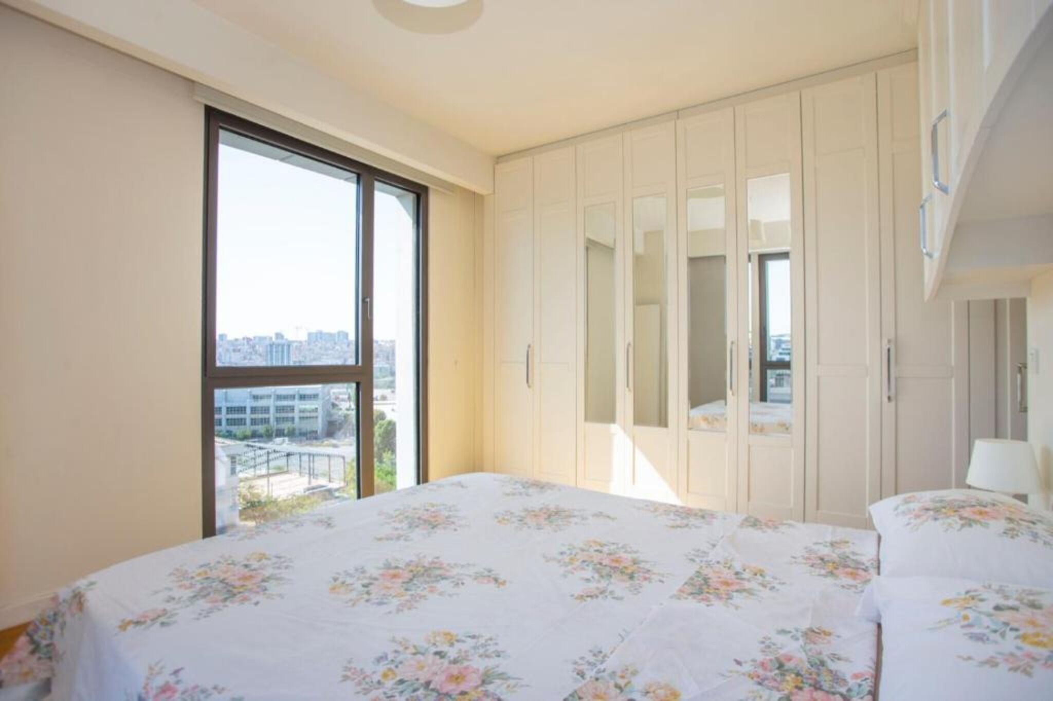 A bedroom with a large window, a bed with floral bedding, and a wardrobe.