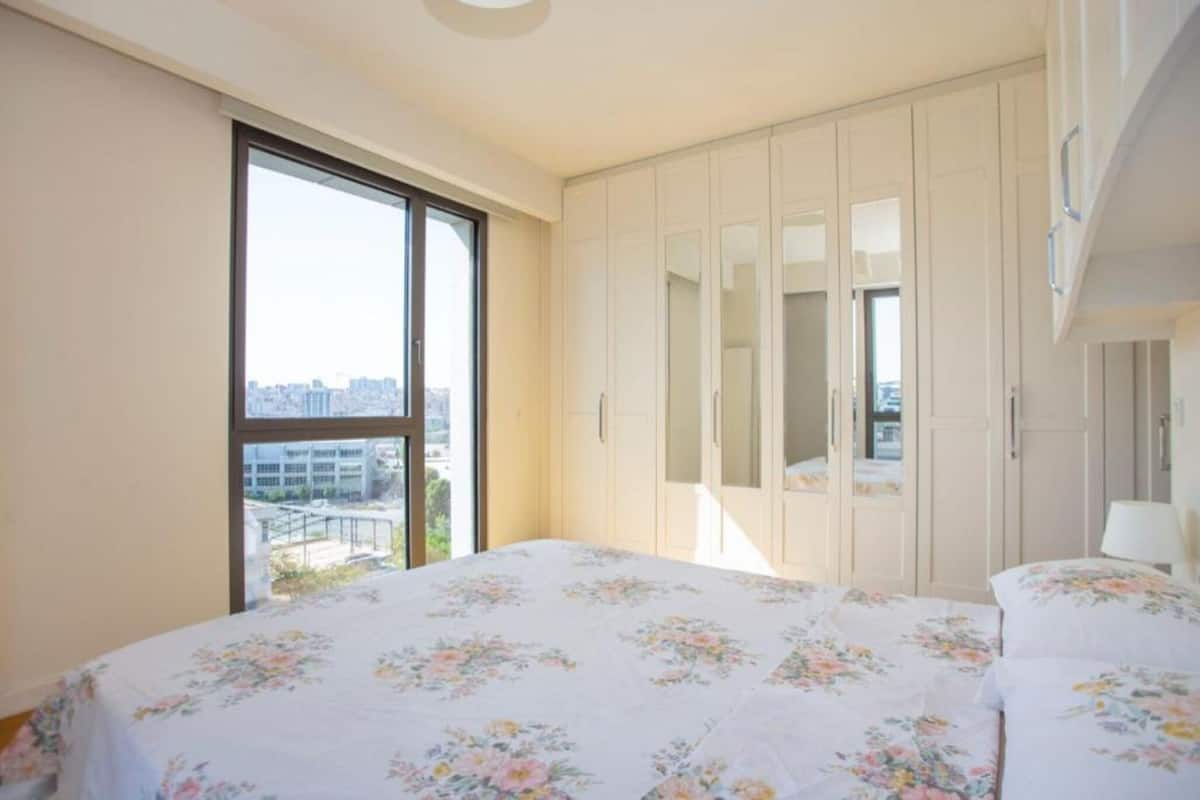 A bedroom with a large window, a bed with floral bedding, and a wardrobe.