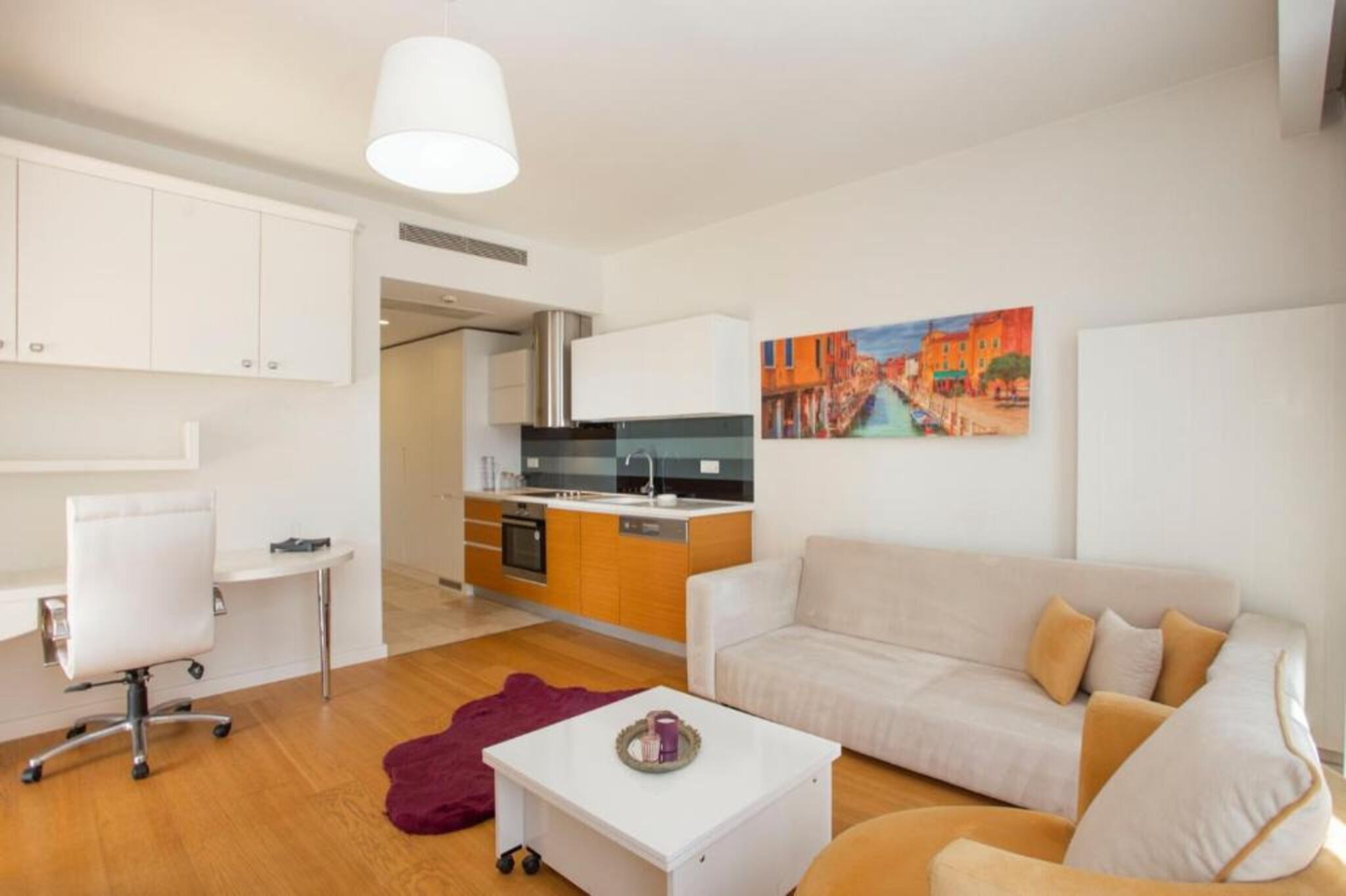 A modern living room with a sofa, a coffee table, a desk, and a kitchen area.