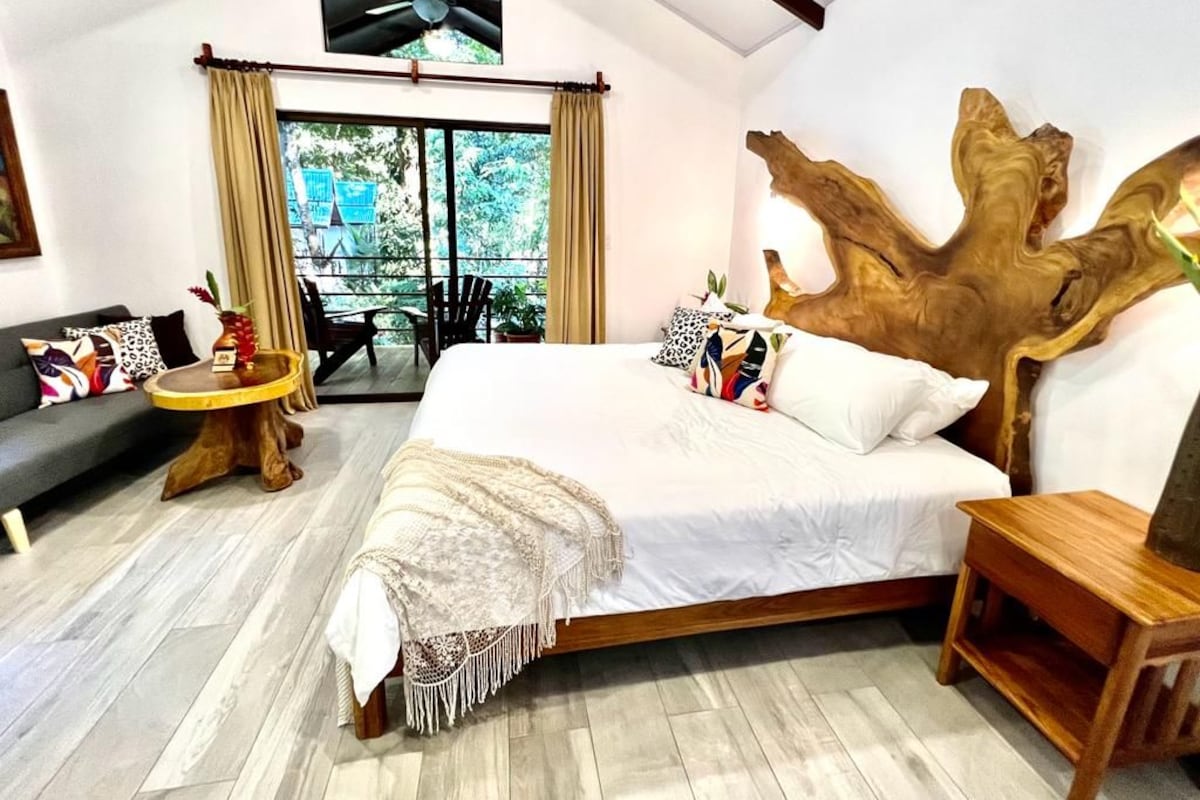 A bedroom with a large bed, a wooden headboard, a sofa, a small round table, and a balcony with a view.