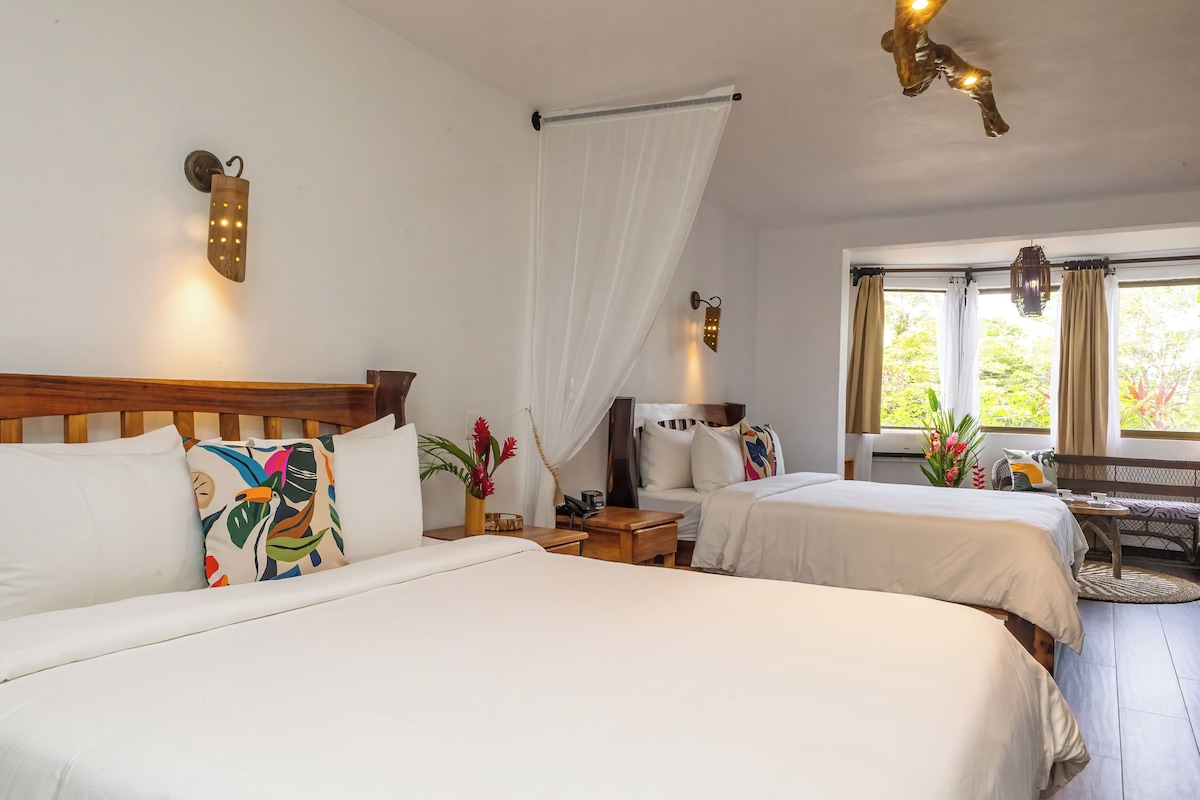 A hotel room with two beds, a wooden headboard, a bedside table with a lamp, a window with curtains, and a view of greenery outside.