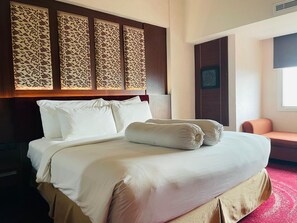 A hotel room with a bed, pillows, a headboard with a patterned design, a chair, and a window. - I Lux Hotel Jogja (Ngaglik)