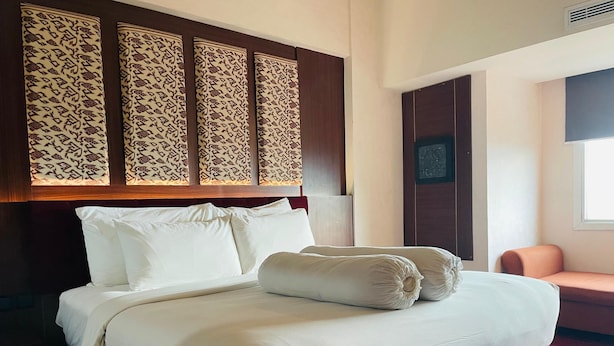 A hotel room with a bed, pillows, a headboard with a patterned design, a chair, and a window. - I Lux Hotel Jogja (Ngaglik)