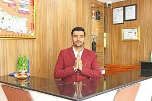 A man in a red suit standing at a reception desk with a wooden wall, certificates, and a map in the background. - Hotel Aaradhya (Udaipur)