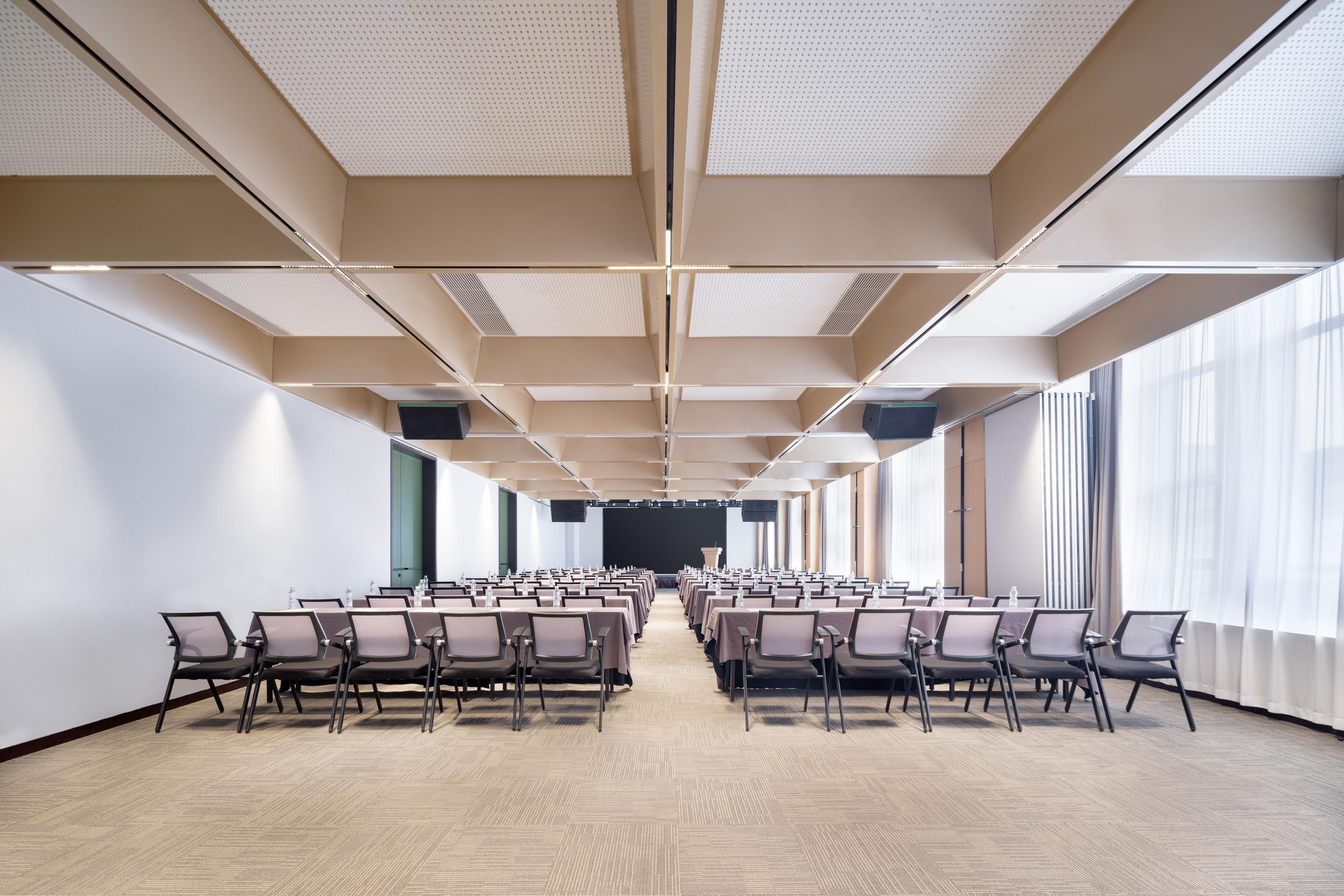 A conference room with rows of chairs, a stage at the front, and large windows on one side.