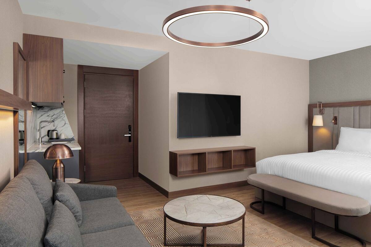 Ein modernes Hotelzimmer mit einer Couch, einem runden Couchtisch, einem Bett, einem Fernseher, einer Bank, einem Holzschrank und einer Deckenleuchte.