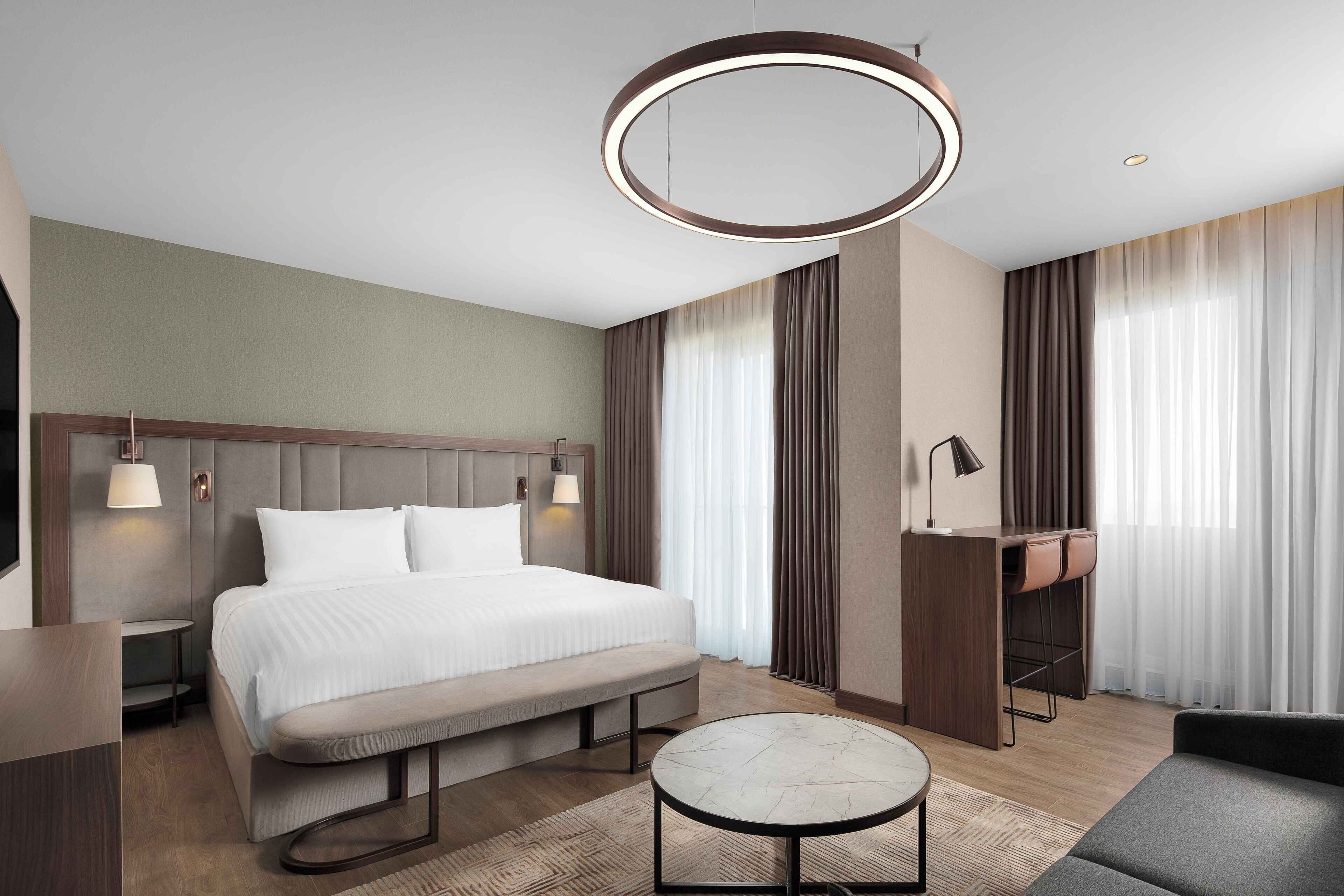 A modern hotel room with a bed, a desk, a bench, a round table, and a hanging light fixture.