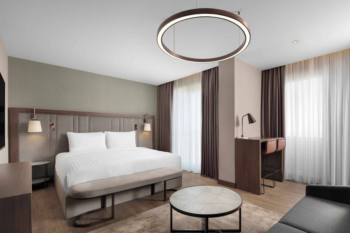 Ein modernes Hotelzimmer mit Bett, Schreibtisch, einer Bank, einem runden Tisch und einer Pendelleuchte.