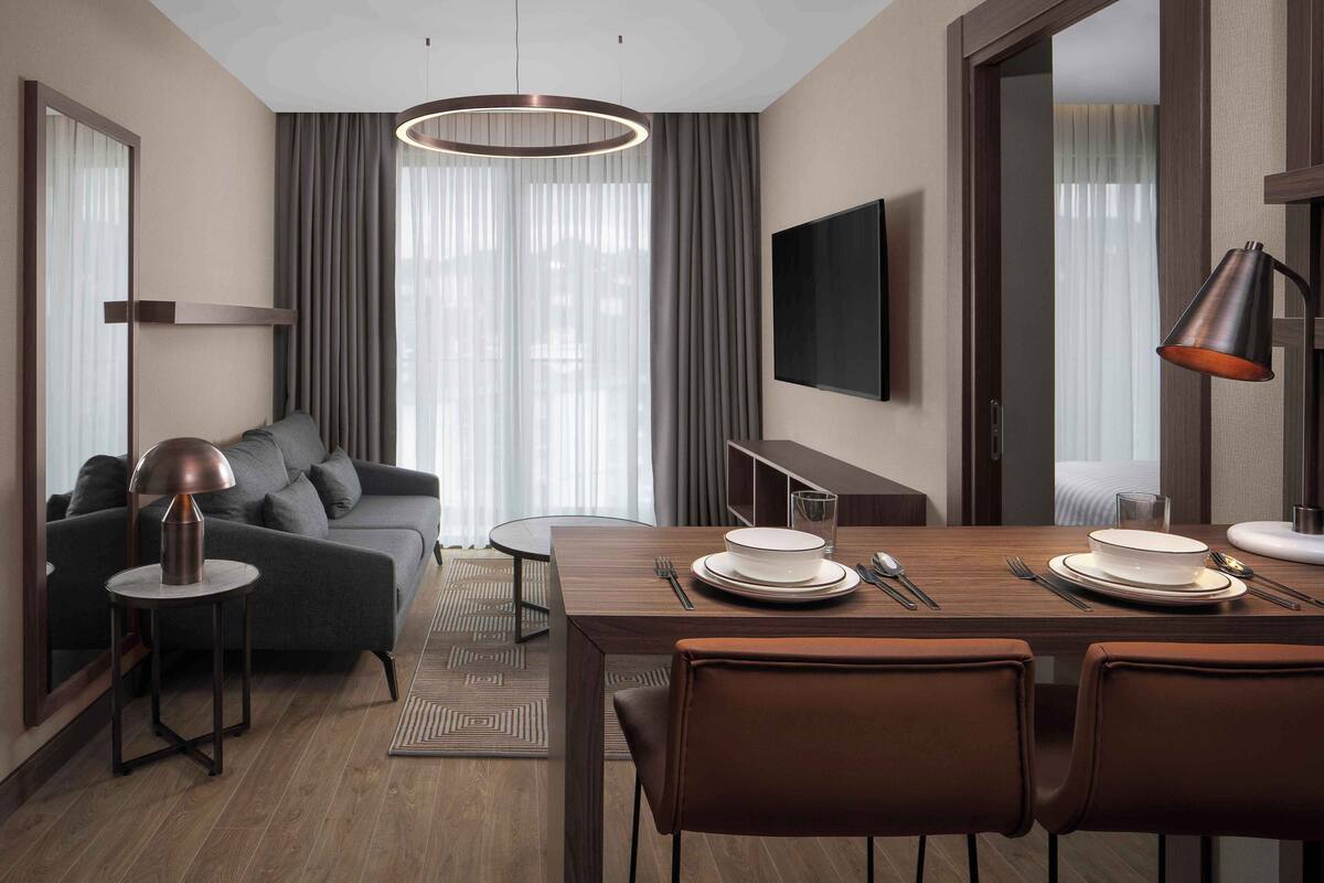 Ein modernes Hotelzimmer mit Essbereich, Sofa, Fernseher und einem großen Fenster mit Vorhängen.