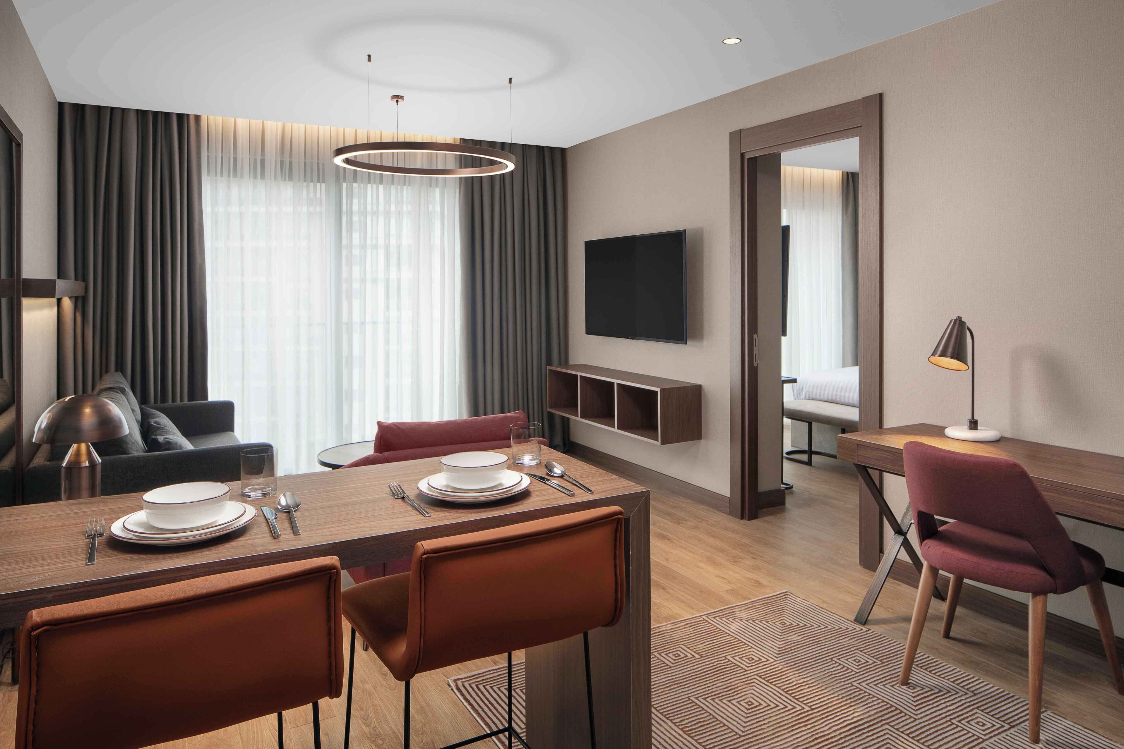 A modern hotel room with a dining area, a TV mounted on the wall, and a view of the cityscape through the window.