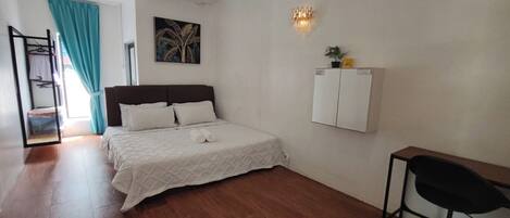 Un dormitorio con cama, escritorio, silla, ventilador de techo, mueble de pared y un cuadro.
