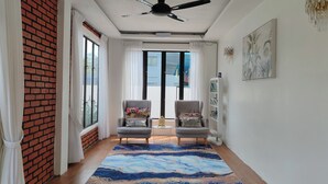 Una habitación con dos sillones, un ventilador de techo, un ventanal con cortinas, una alfombra azul estampada y una estantería en la pared.