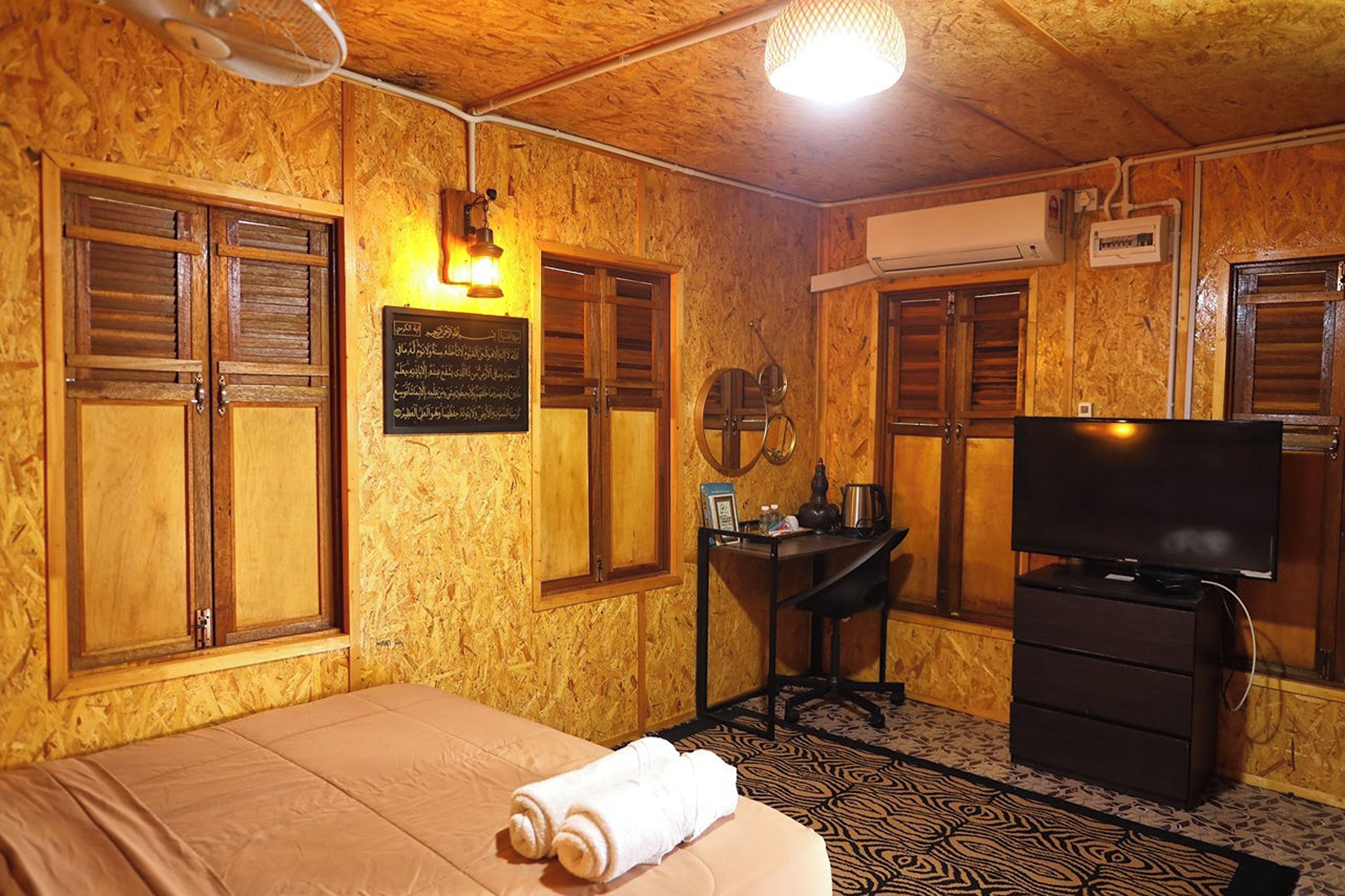 A room with wooden walls, a bed with a beige bedspread, a desk with a chair, a television, and a patterned rug.