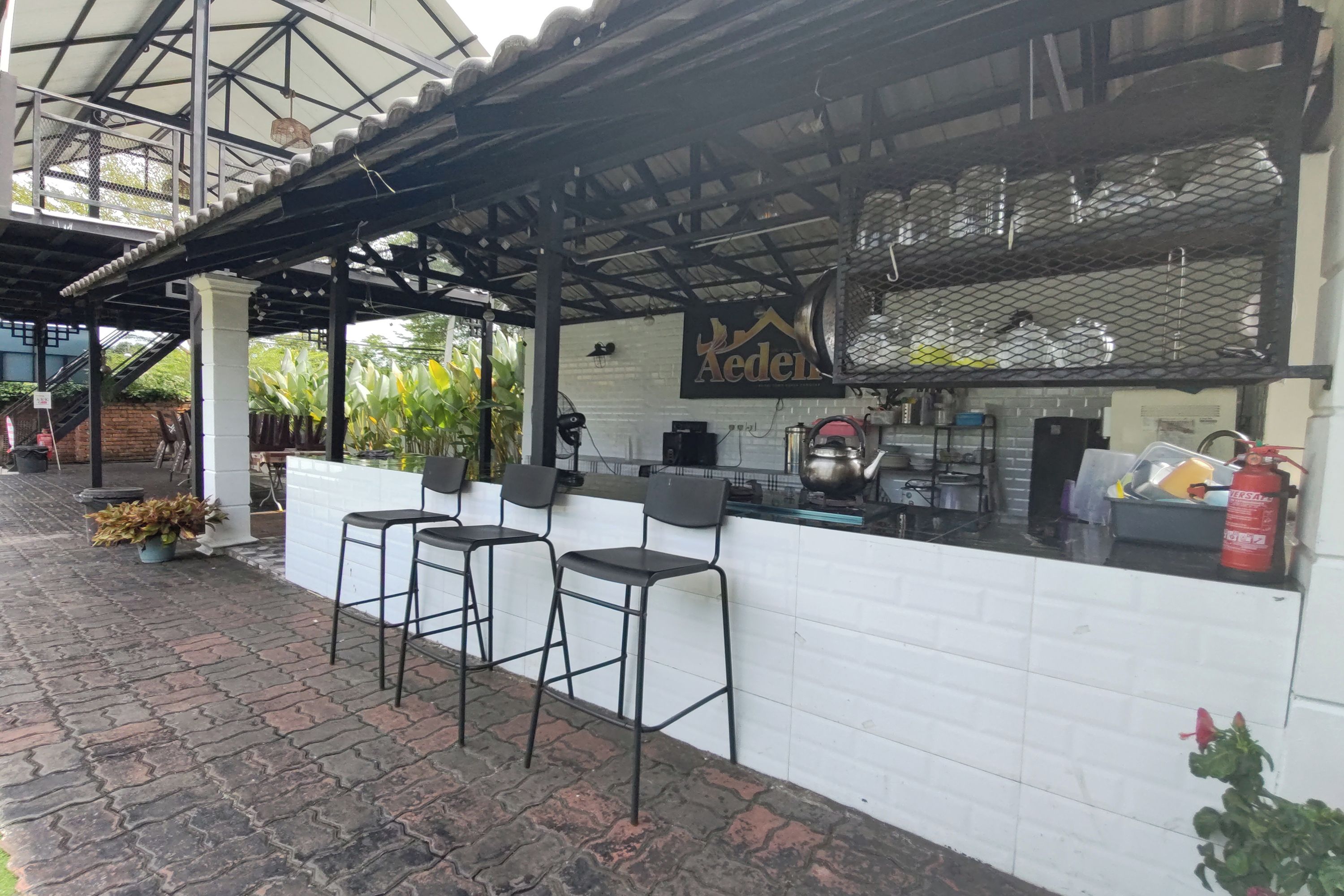 A restaurant with a white tiled exterior, a metal roof, and a sign that reads 'Aeden.' There are three black bar stools and a counter with kitchen equipment.