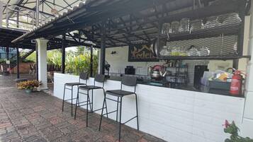 A restaurant with a white tiled exterior, a metal roof, and a sign that reads 'Aeden.' There are three black bar stools and a counter with kitchen equipment.