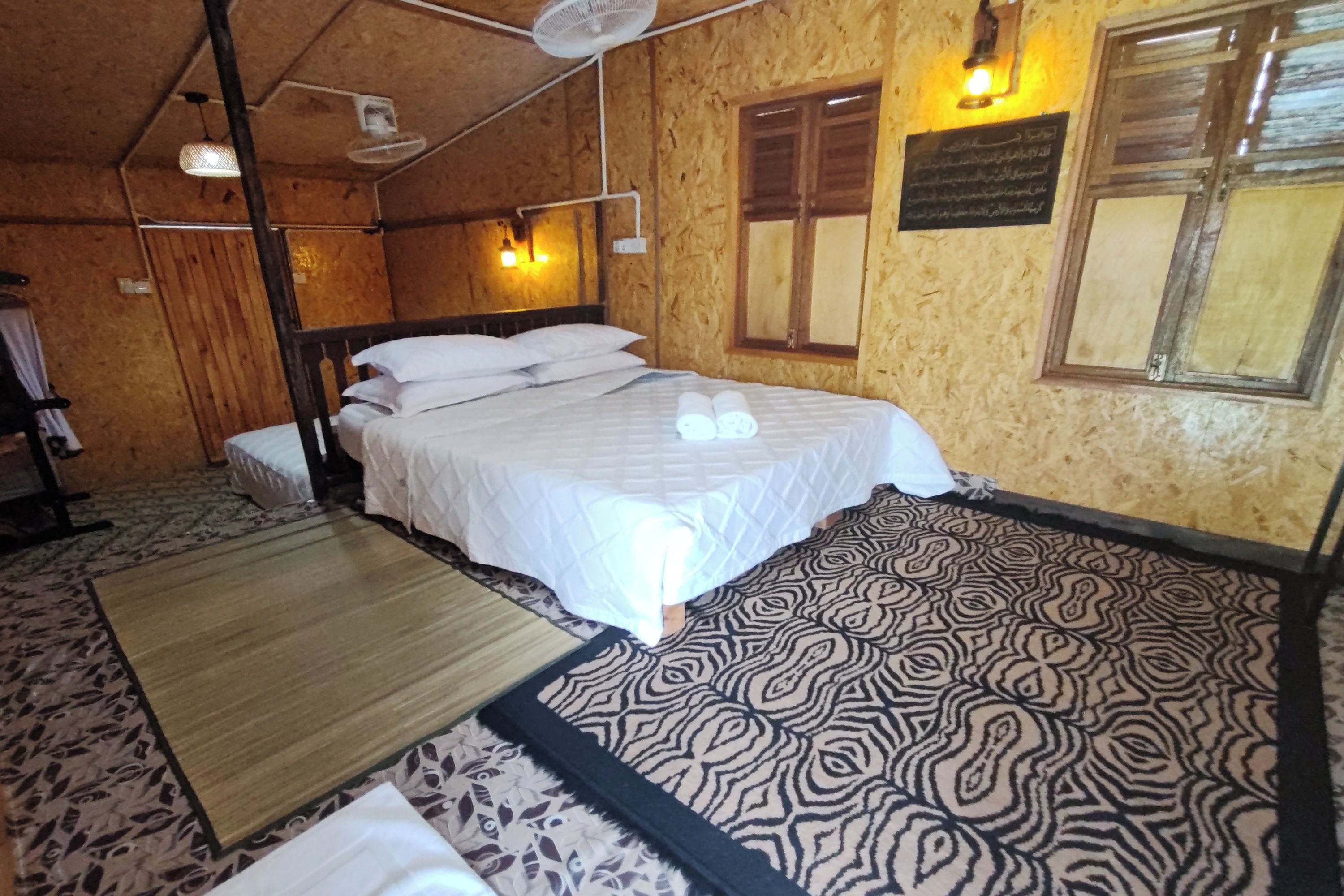 A room with a bed, a fan, a window, and a wall-mounted light fixture.