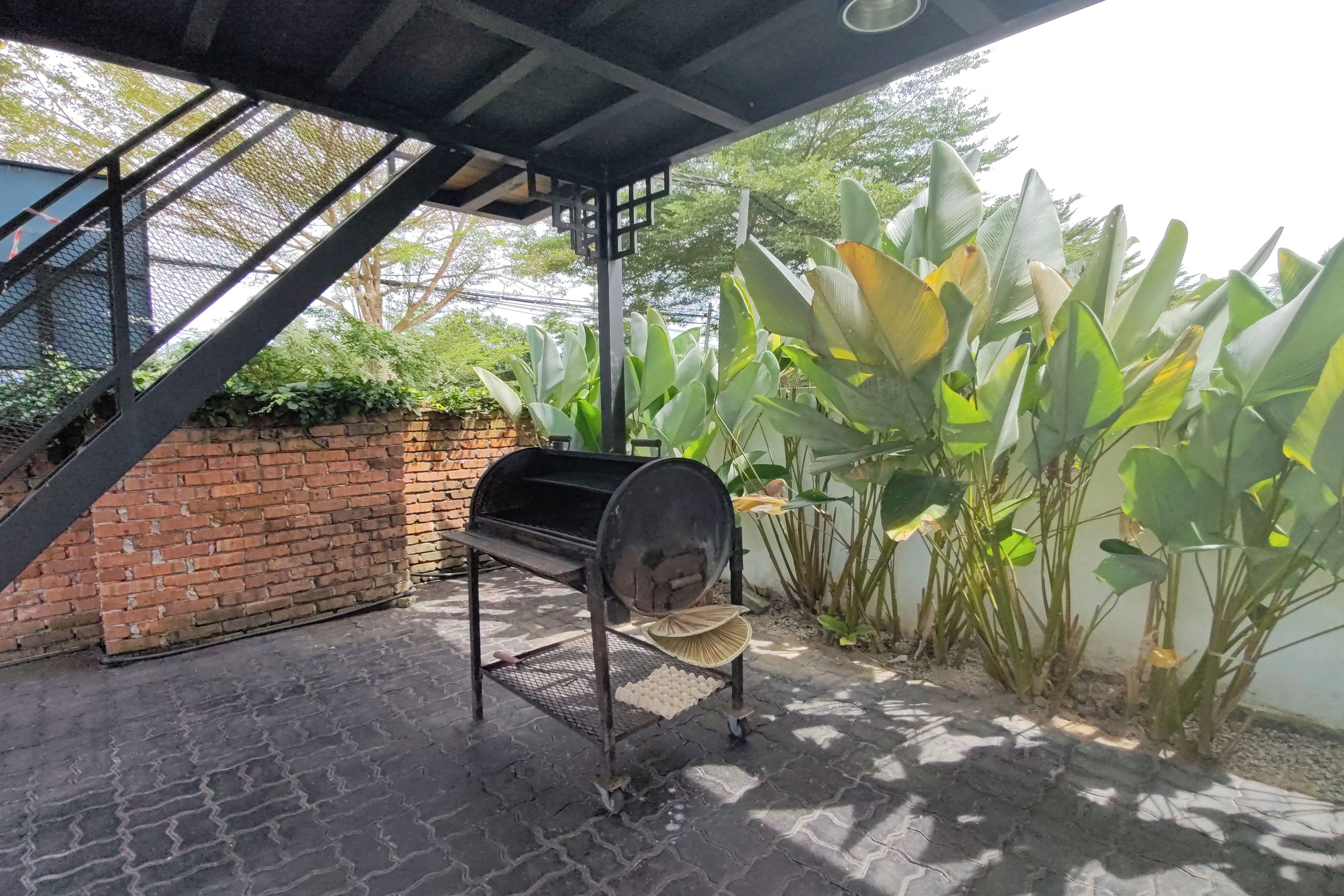 A barbecue grill under a covered patio area with a brick wall and large leafy plants.