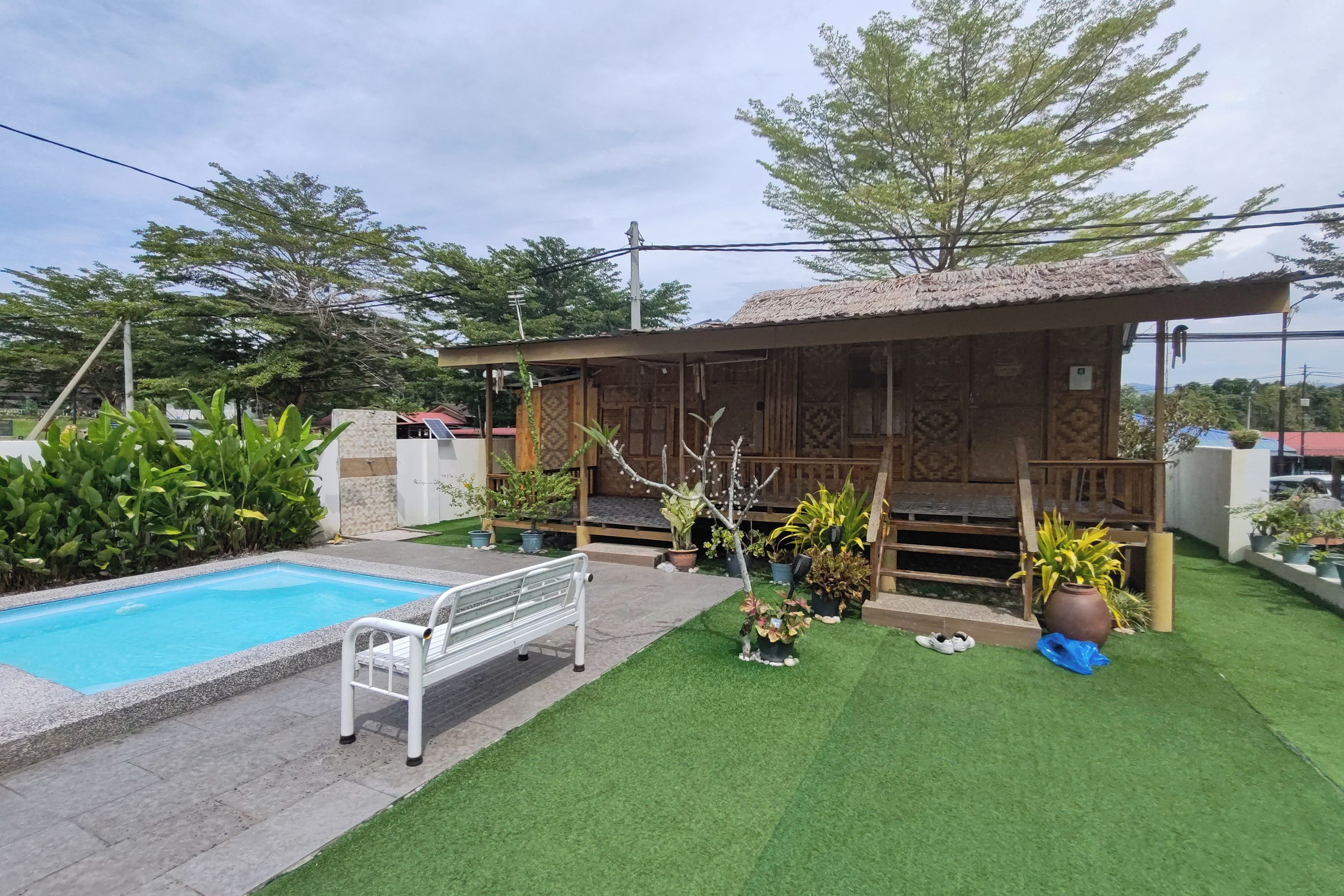 A pool with a white bench, a wooden structure with a thatched roof, and various potted plants.
