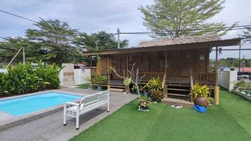 A pool with a white bench, a wooden structure with a thatched roof, and various potted plants.