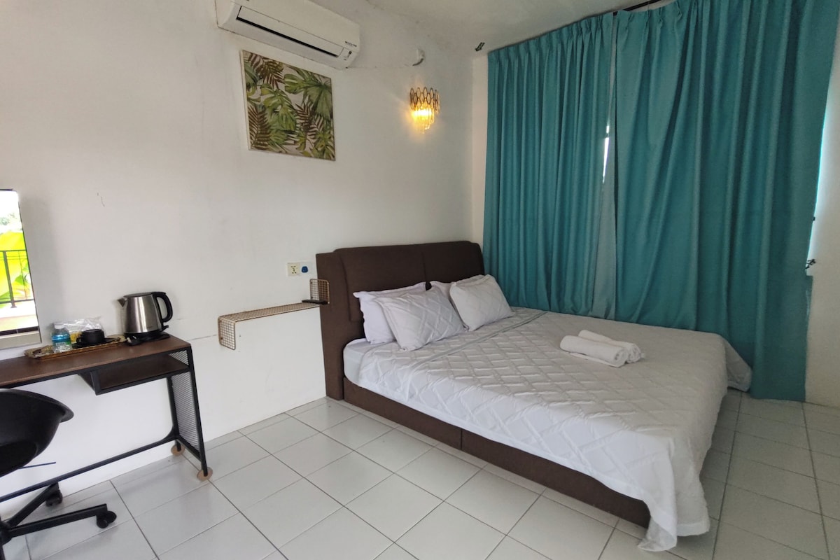 A hotel room with a bed, a desk with a chair, a wall-mounted air conditioner, a wall-mounted light fixture, and a window with curtains.