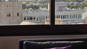 Une chambre avec vue sur le paysage urbain, où l’on peut apercevoir un bâtiment portant l’inscription « HELLENIC BANK ».