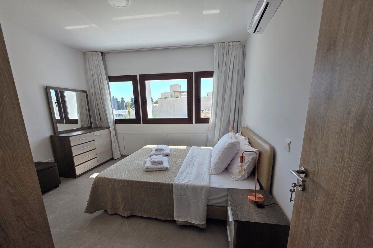 A modern bedroom with a large bed, a bedside table, a dresser, and a mirror.