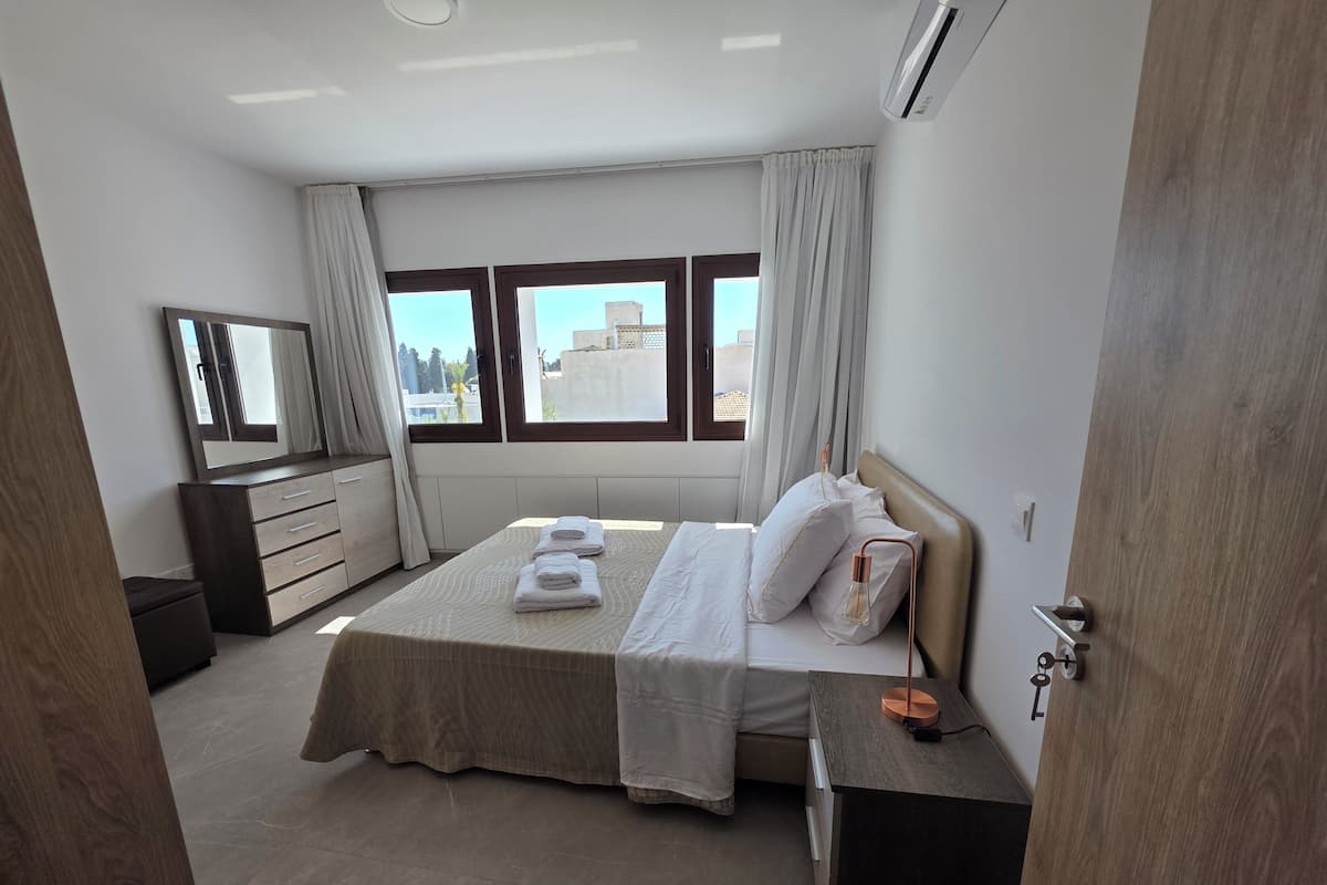 A modern bedroom with a large bed, a bedside table, a dresser, and a mirror.