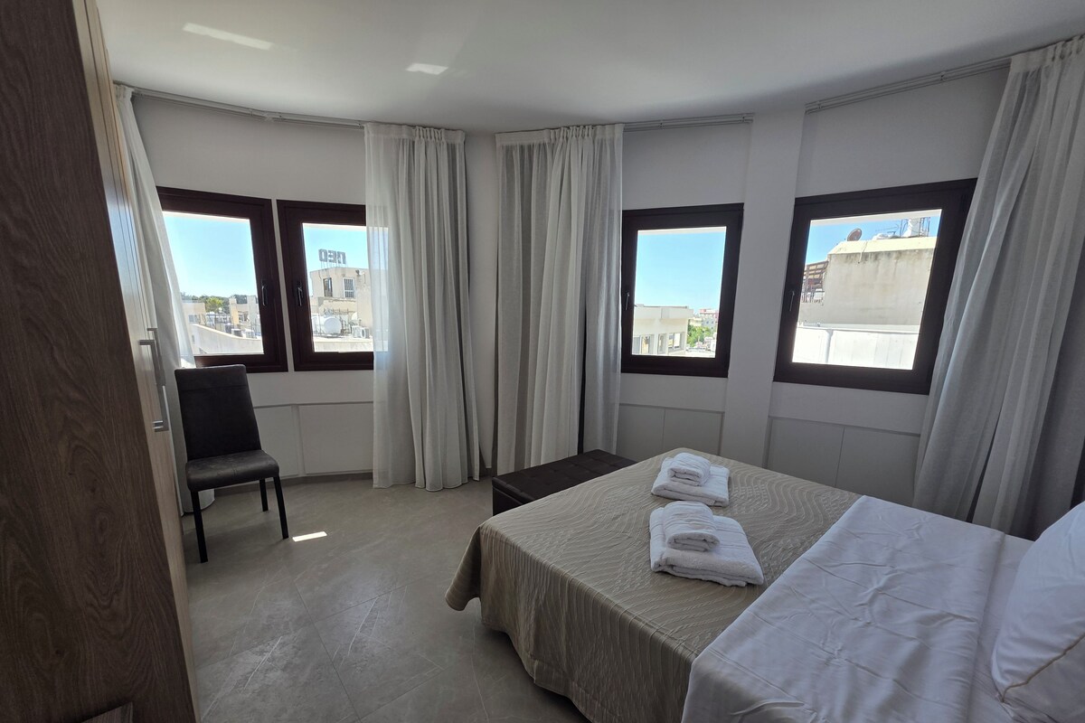 A hotel room with a bed, a chair, and two windows with curtains.