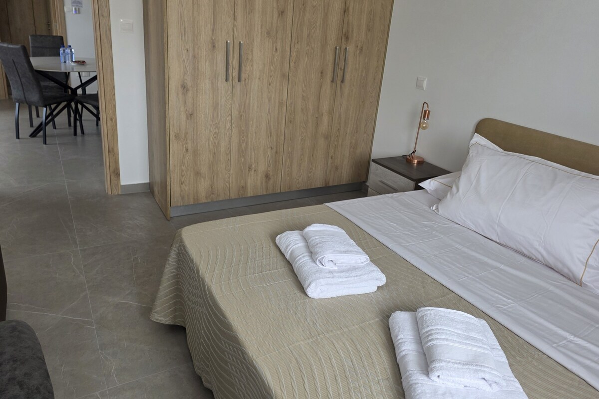 A hotel room with a bed, a wooden wardrobe, a desk with chairs, and a nightstand with a lamp.