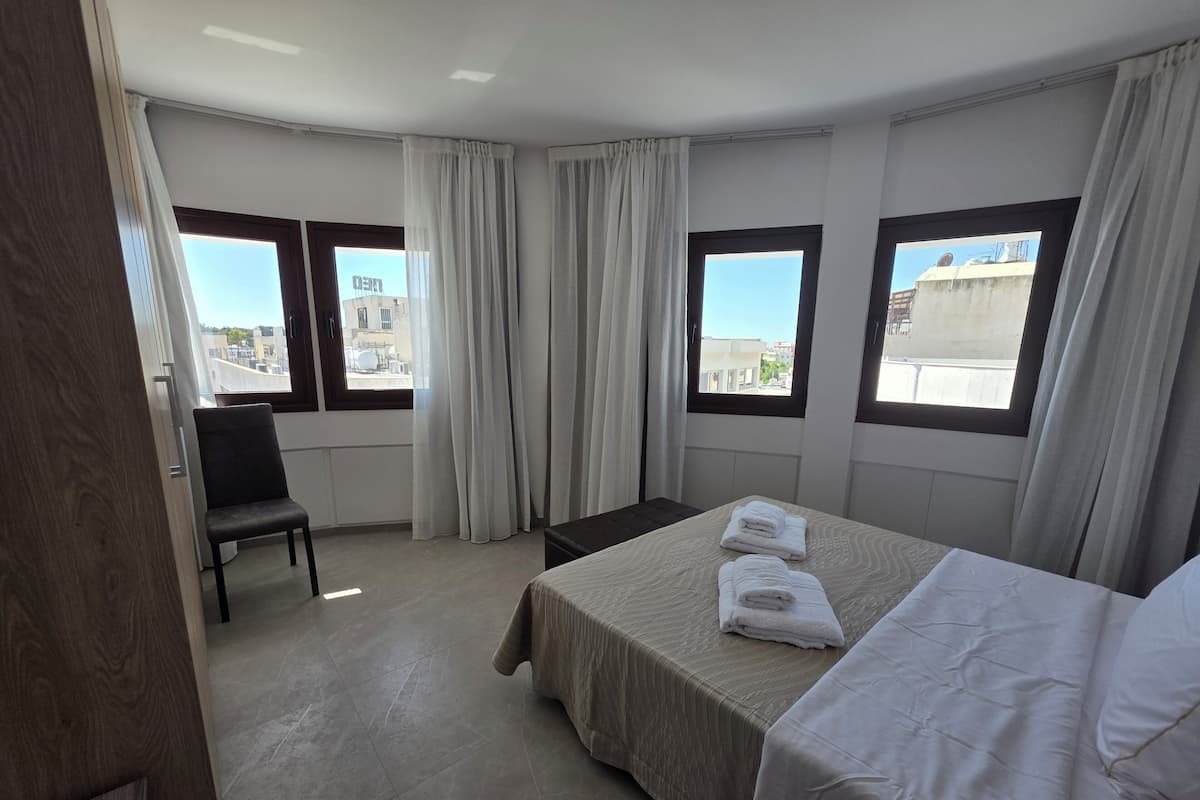 A hotel room with a bed, a chair, and two windows with curtains.
