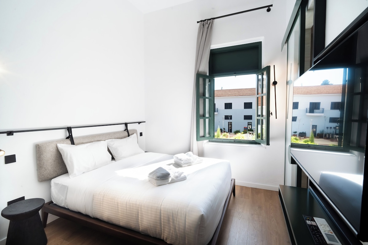 A modern hotel room with a bed, a small table, a TV, and a window with a view of buildings.