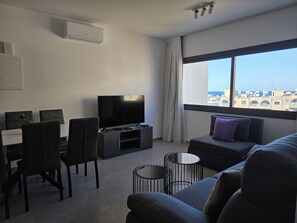 A modern living room with a flat-screen TV, grey sofas, a dining table with chairs, and a large window with a city view. - Kentrikon 1924 Boutique Hotel And Suites (Paphos)