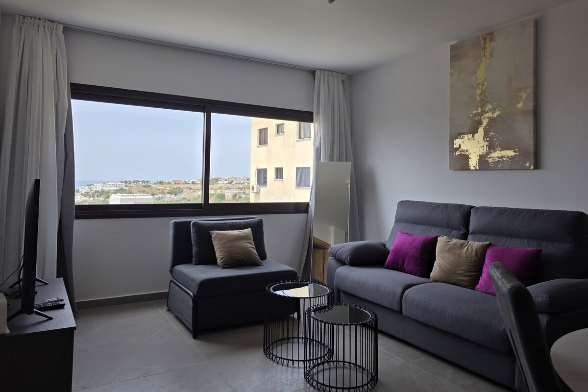 A modern living room with a grey sofa, a black armchair, and a round coffee table. A large window offers a view of buildings and greenery.